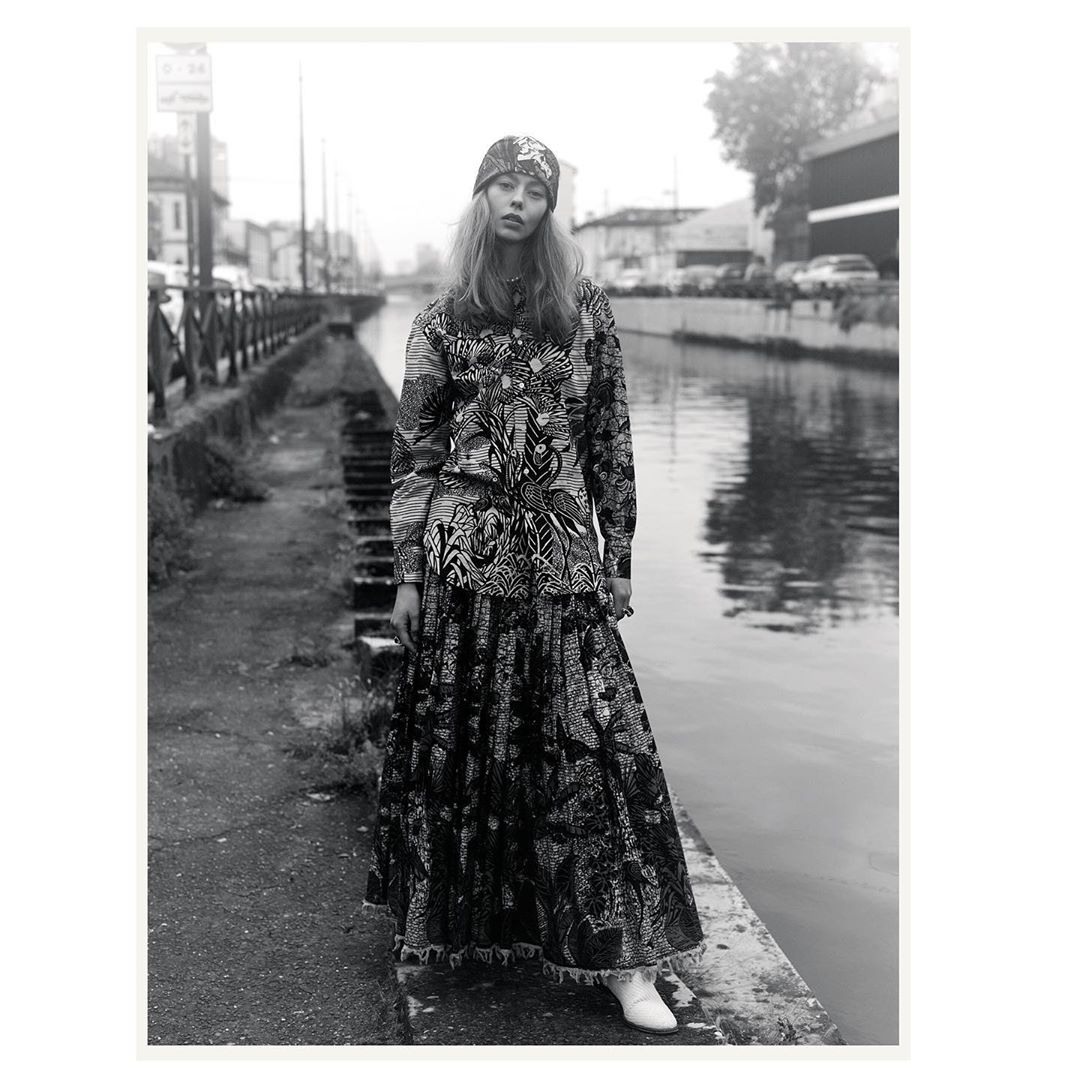 Fashion photo of a young adult female model posing outdoors wearing a patterned headscarf, top, and long skirt with white ankle boots.