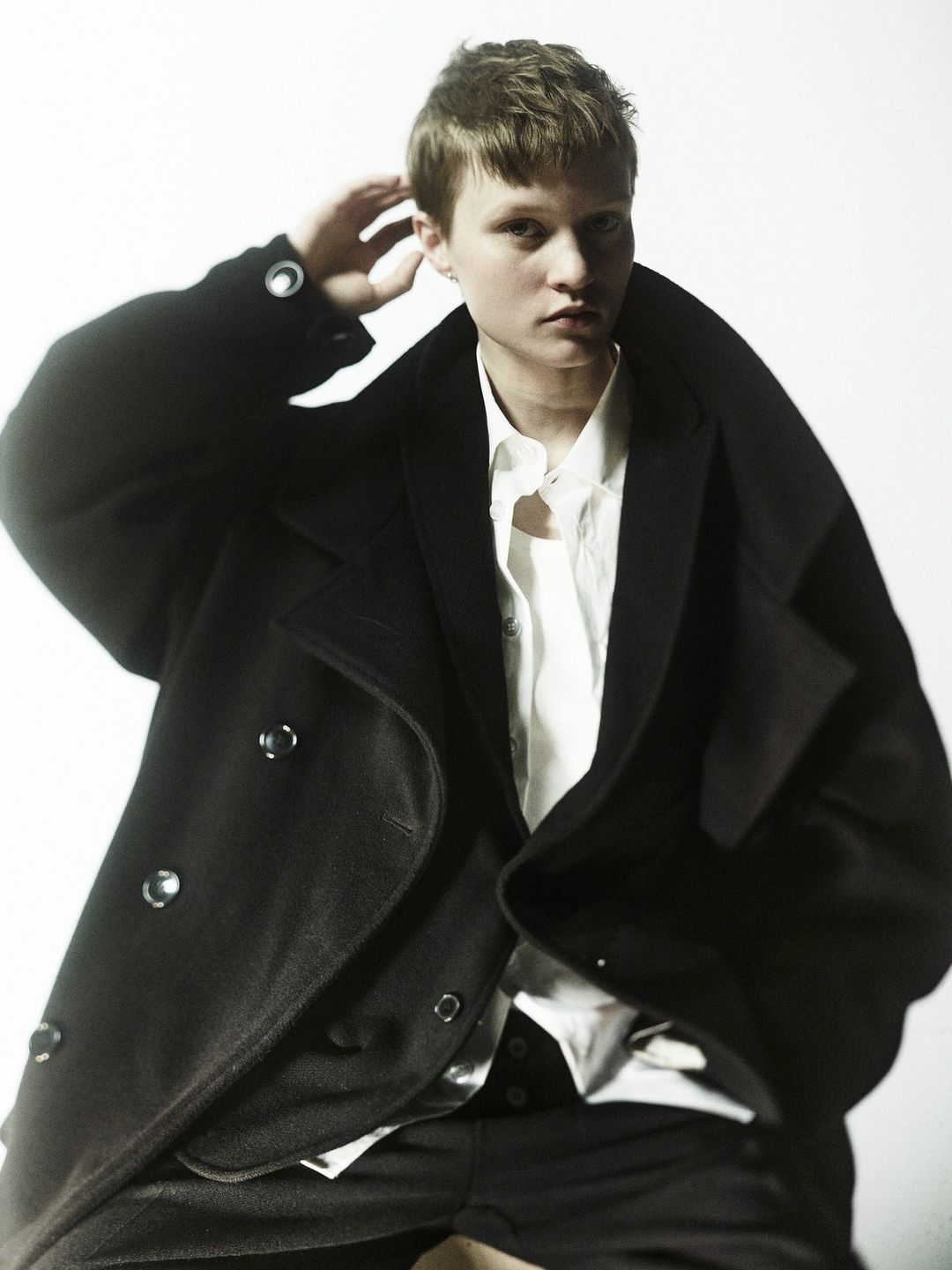 Fashion portrait of a person with short hair wearing a black coat and white shirt against a white background in a studio setting.