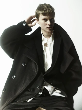 Fashion portrait of a person with short hair wearing a black coat and white shirt against a white background in a studio setting.