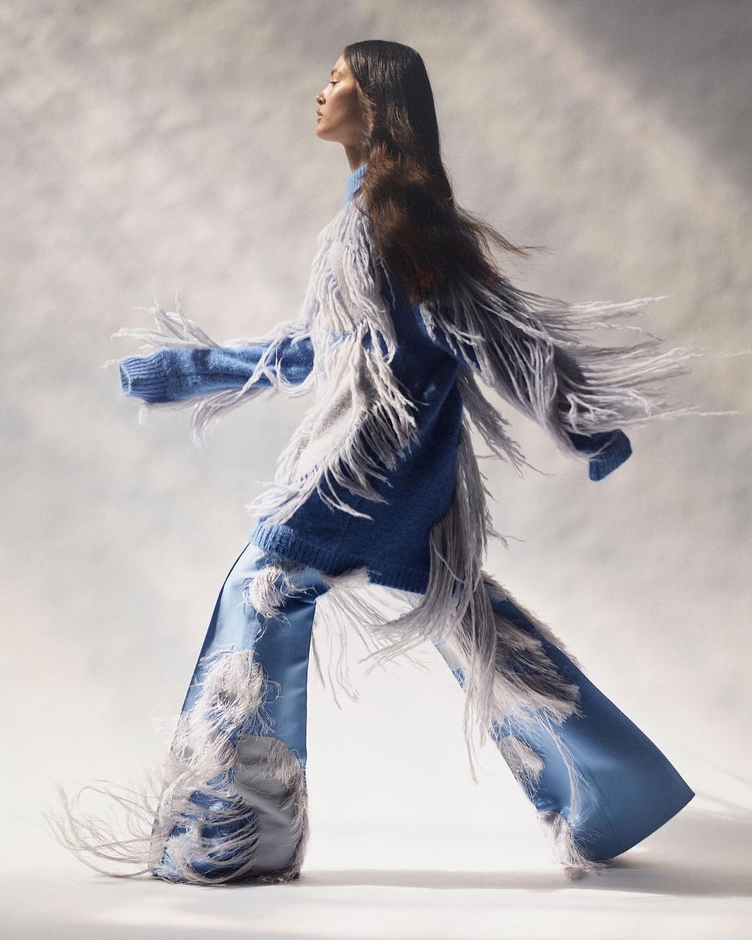 Woman in a fringed blue knit sweater and wide-leg satin pants walks against a soft, diffused background in a fashion-style studio shot.