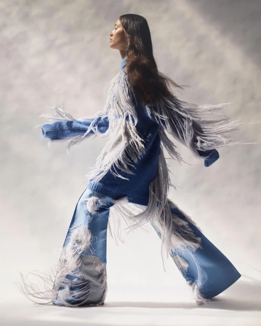 Woman in a fringed blue knit sweater and wide-leg satin pants walks against a soft, diffused background in a fashion-style studio shot.
