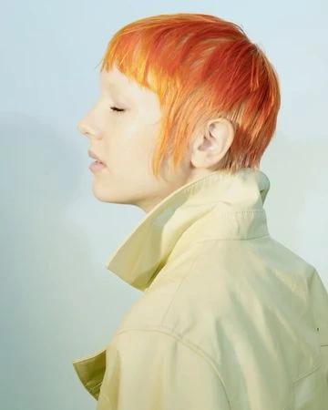 Profile view of an adult female with orange short hair and a light skin tone wearing a light-yellow coat against a pale-blue background.