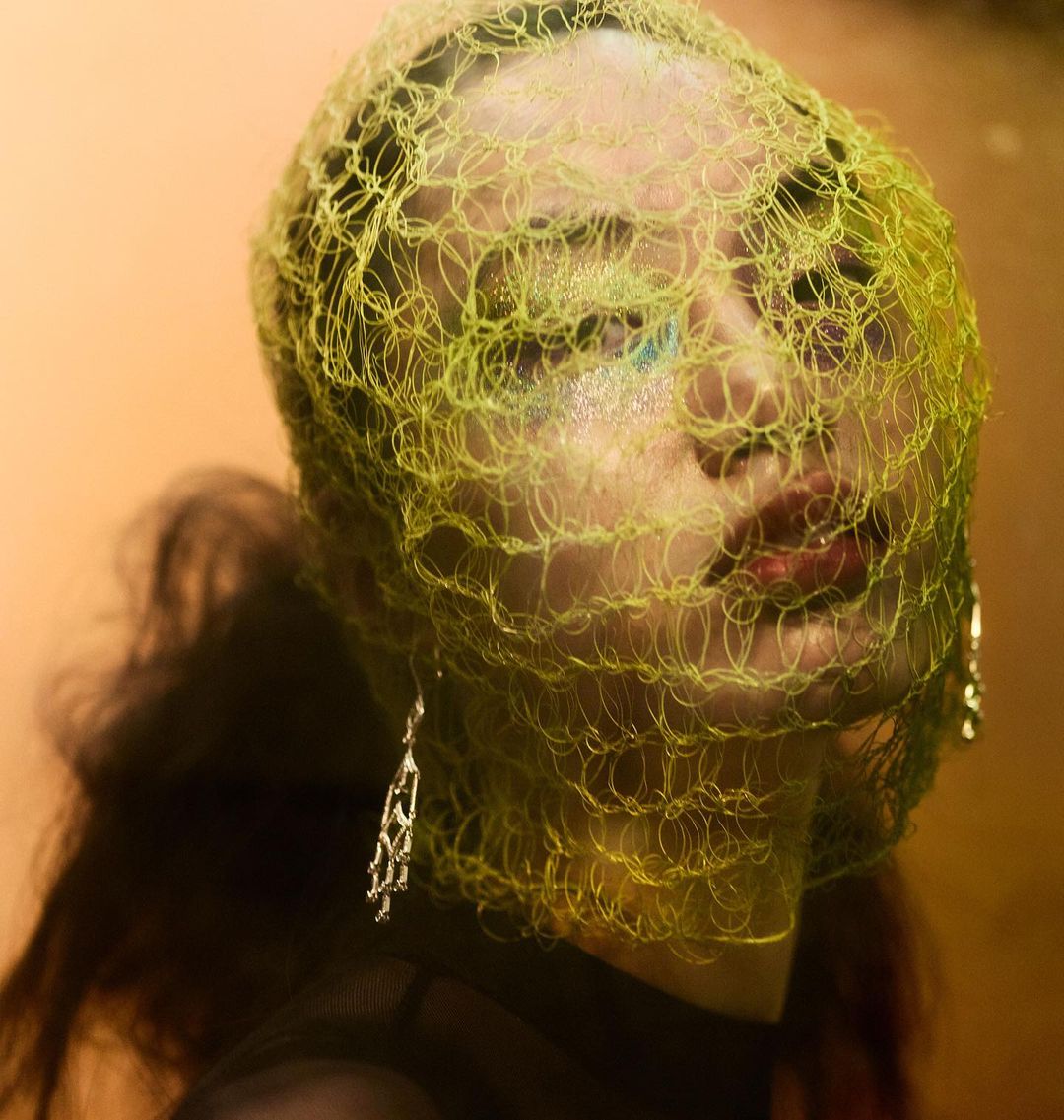 Close-up portrait of an adult female with light skin, wearing a yellow net head covering and silver dangle earrings against an orange background.
