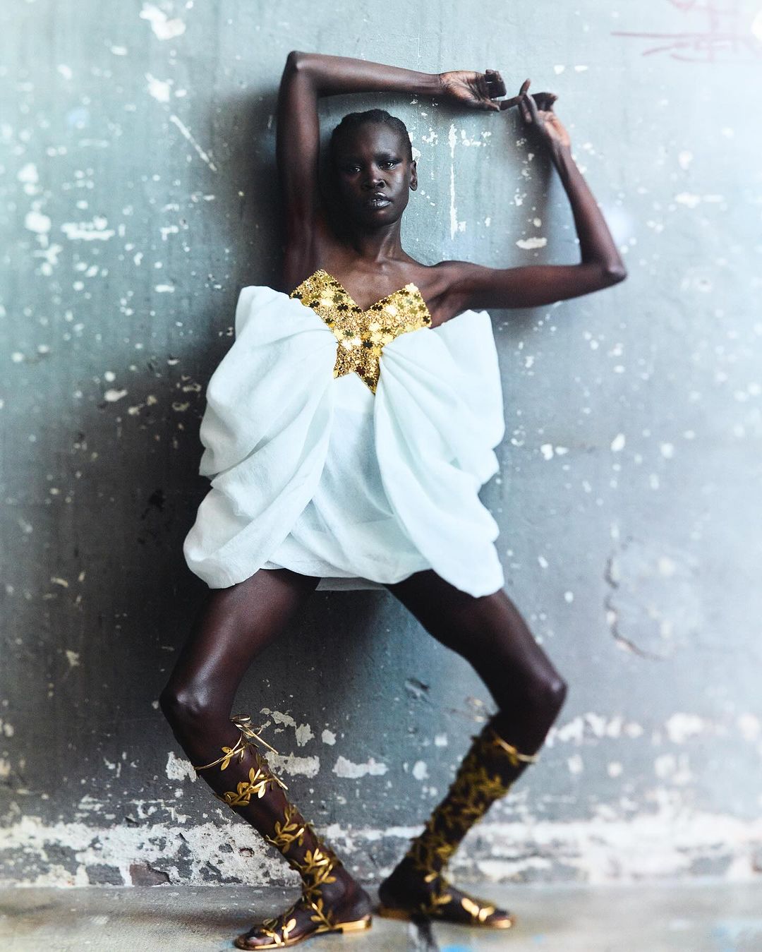 Adult woman in a white dress with gold accent and gold boots, poses against a textured wall. Fashion editorial image, studio lighting.
