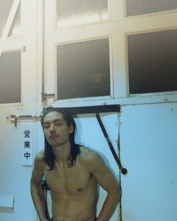 Shirtless adult male with long hair poses indoors against a white wall with a window, looking towards the camera with a neutral expression.