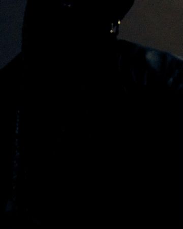 Dark, low-light image of an adult with a hat and hooded jacket partially visible; the person is obscured by shadows.