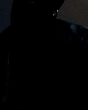 Dark, low-light image of an adult with a hat and hooded jacket partially visible; the person is obscured by shadows.