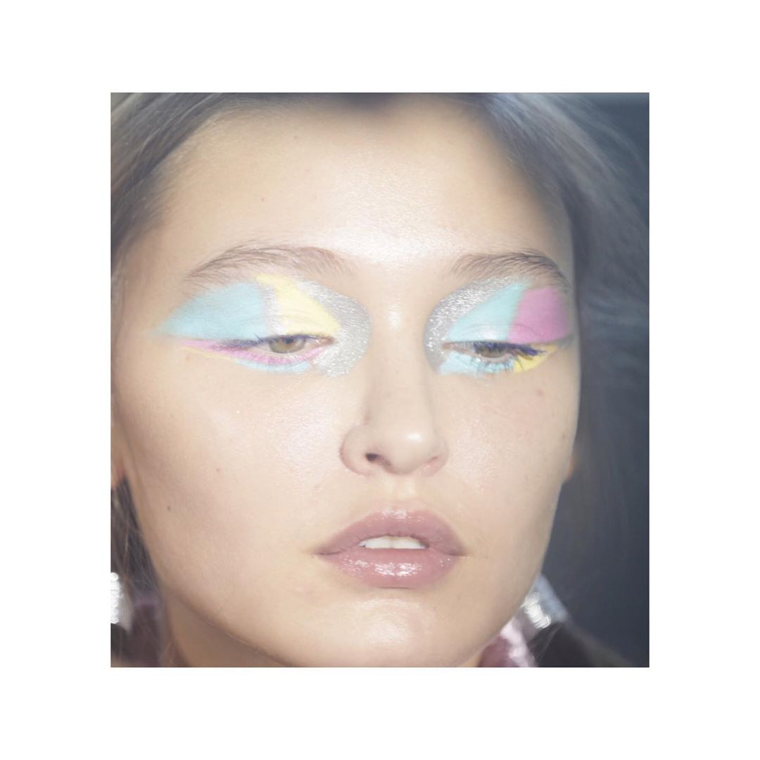 Close-up of a woman's face with artistic pastel makeup around her eyes and glitter on the brow bone, set against a soft background.
