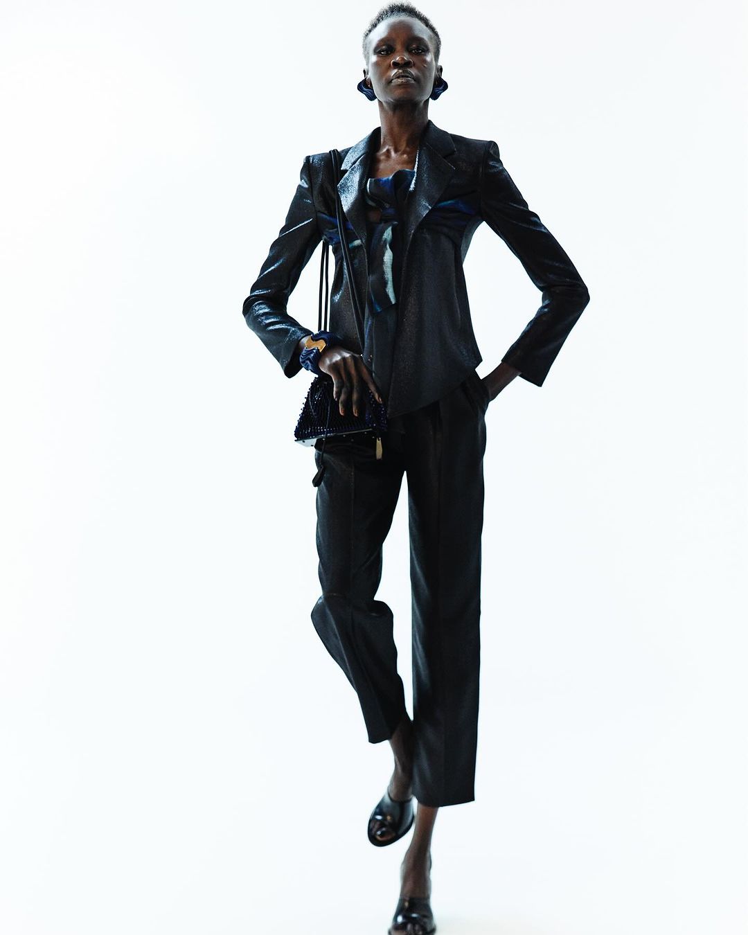 A solo Black fashion model poses confidently in a dark, sleek suit with a small purse against a bright white background in a studio setting.
