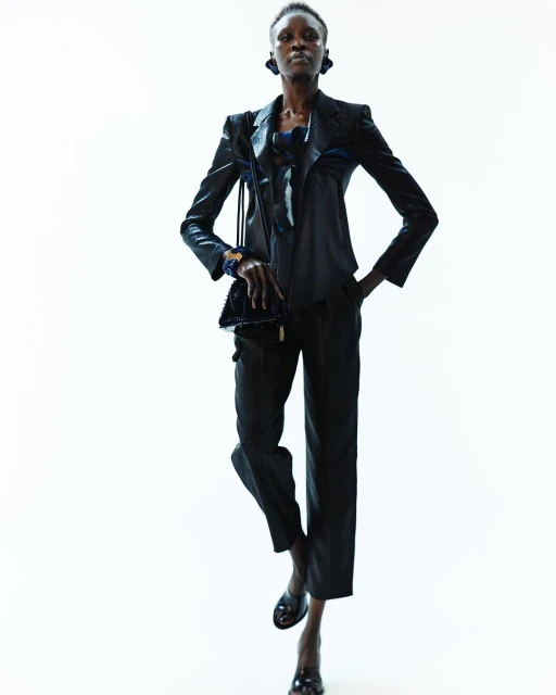 A solo Black fashion model poses confidently in a dark, sleek suit with a small purse against a bright white background in a studio setting.
