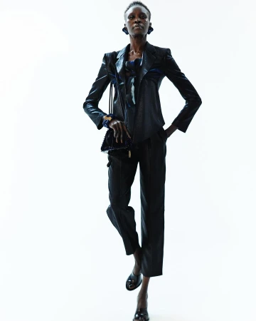 A solo Black fashion model poses confidently in a dark, sleek suit with a small purse against a bright white background in a studio setting.