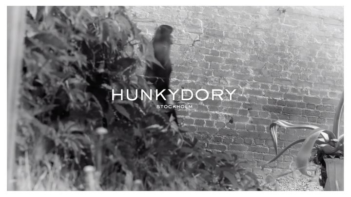Black and white outdoor scene showcases brick wall, greenery, and 'Hunkydory Stockholm' text overlay. Focus on fashion brand aesthetic.