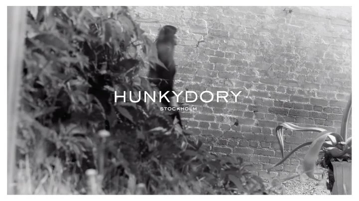 Black and white outdoor scene showcases brick wall, greenery, and 'Hunkydory Stockholm' text overlay. Focus on fashion brand aesthetic.