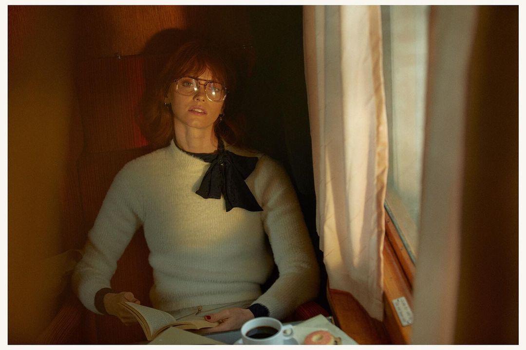 Woman wearing glasses and cream sweater reading a book by a train window with a cup of coffee.