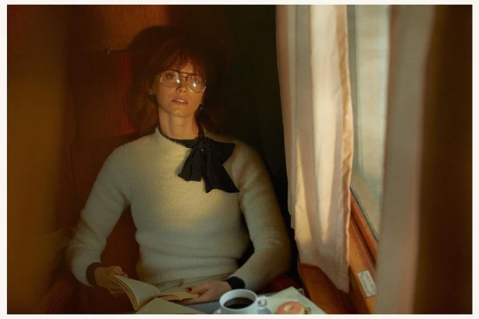 Woman wearing glasses and cream sweater reading a book by a train window with a cup of coffee.