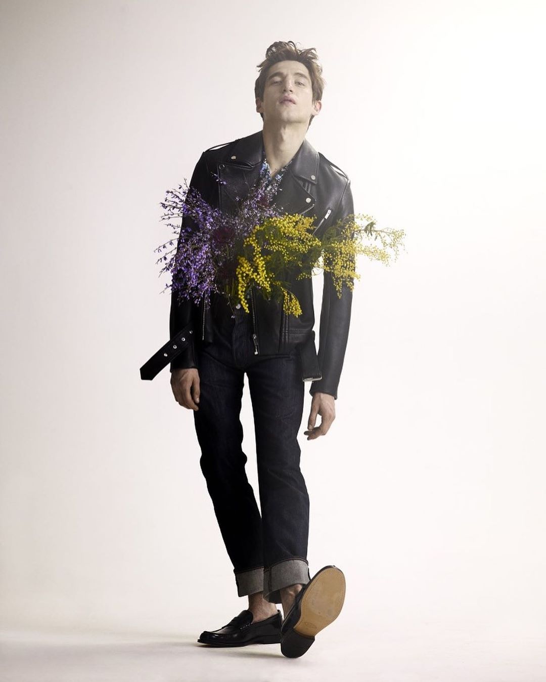 Adult man in a black leather jacket and blue jeans posing with purple and yellow flowers on a white studio background in a fashion shot.