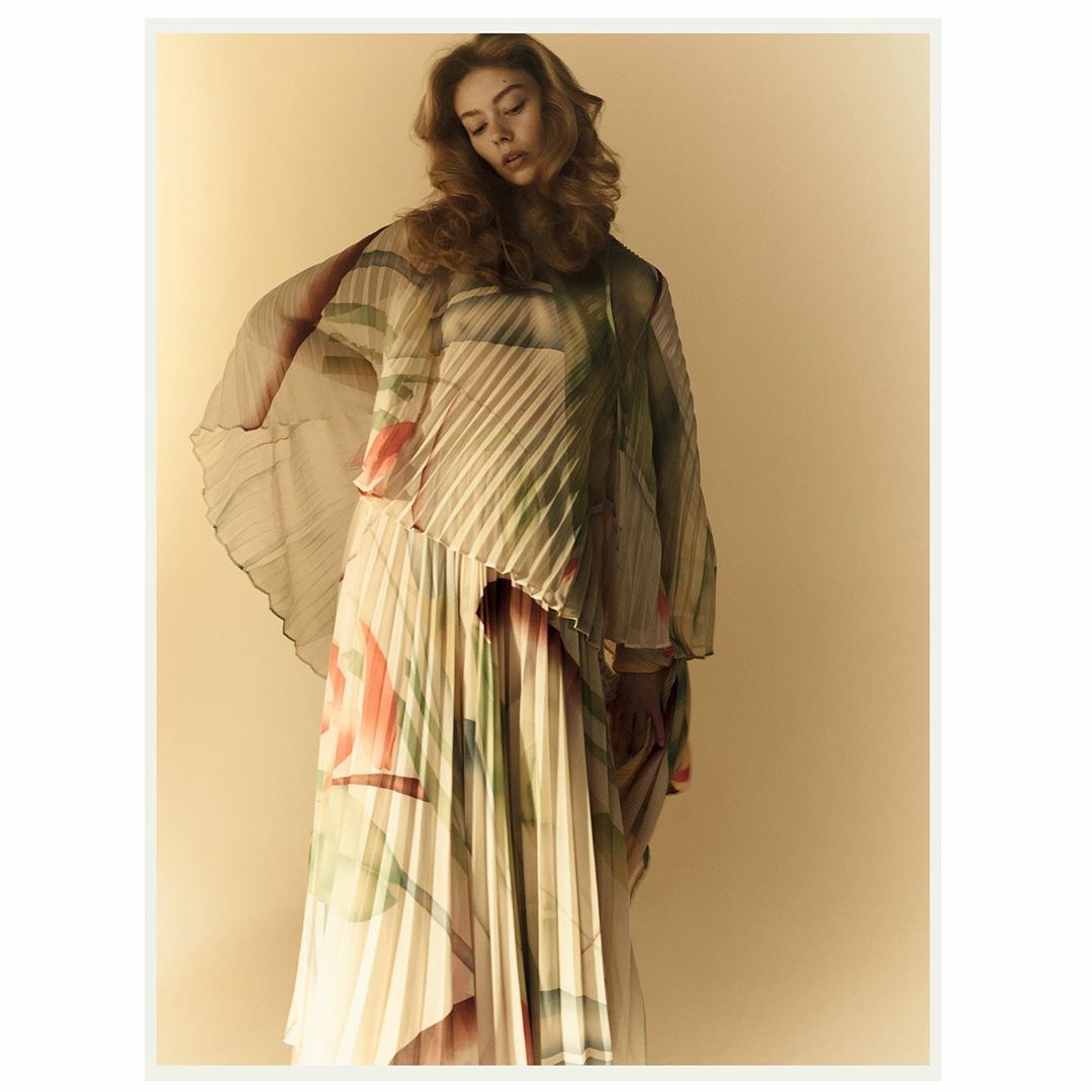 A young woman poses in a flowing, floral-patterned pleated dress against a neutral background in a fashion photography studio.