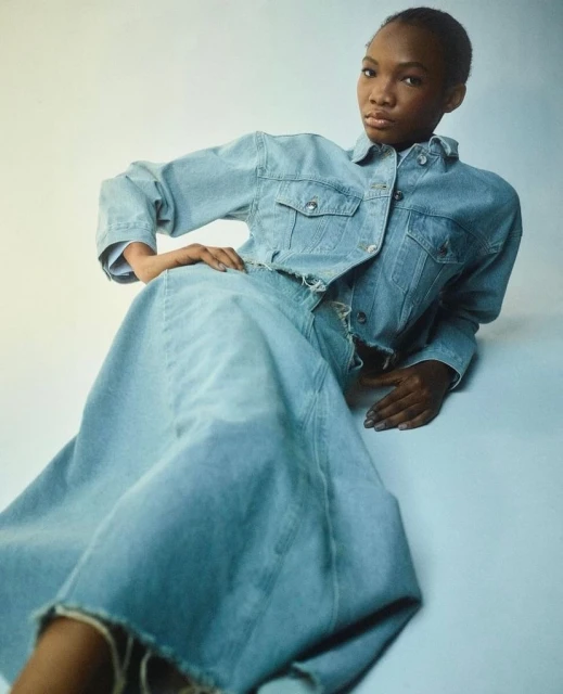 Woman in a light blue denim jacket and matching maxi skirt poses in a studio, fashion shot, simple light background, looking at camera.