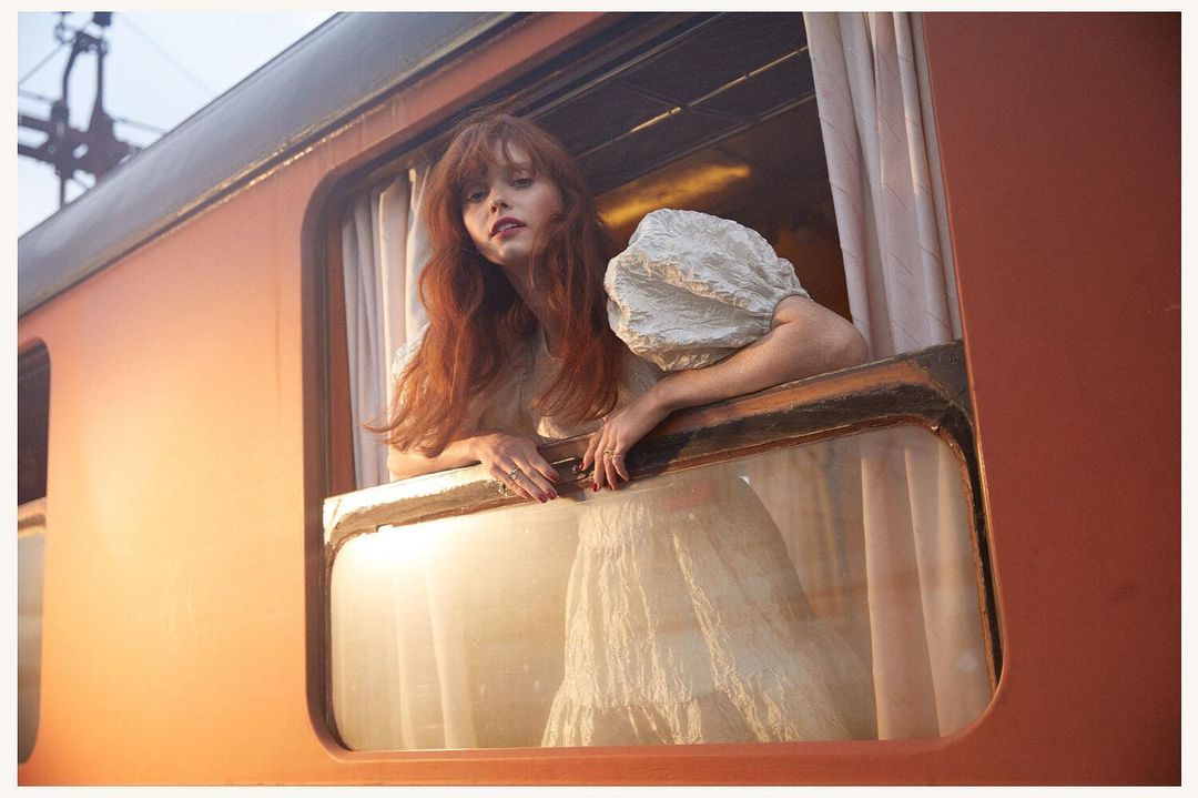 Young woman with red hair in a light dress leans out the window of an orange train car, captured in a soft, dreamy photograph.