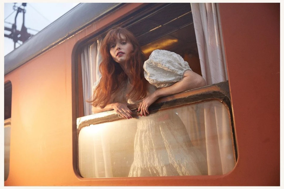 Young woman with red hair in a light dress leans out the window of an orange train car, captured in a soft, dreamy photograph.