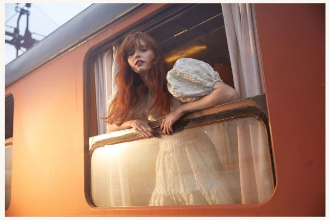 Young woman with red hair in a light dress leans out the window of an orange train car, captured in a soft, dreamy photograph.