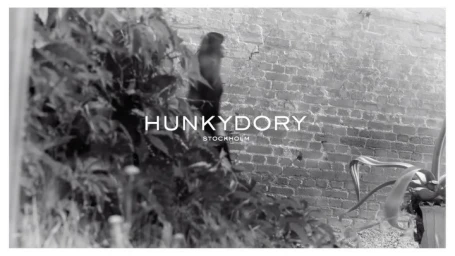 Black and white editorial fashion photograph of a person walking past a brick wall and leafy foliage.