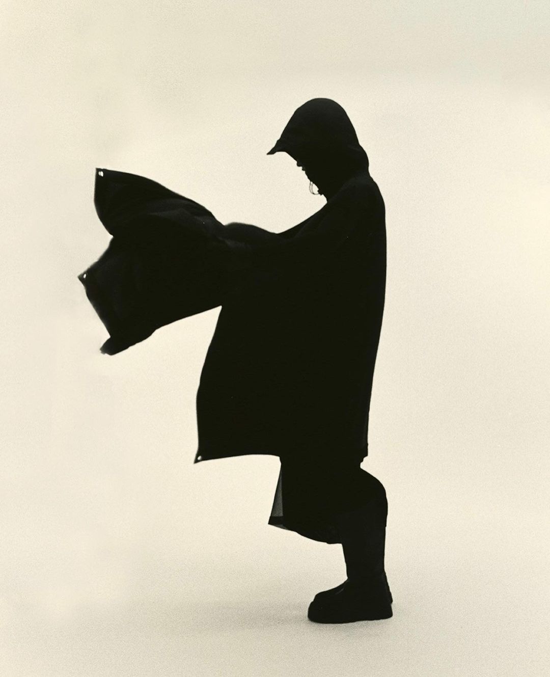A person in a dark hooded raincoat stands on a light surface, holding the coat open with both hands, casting a stark silhouette.
