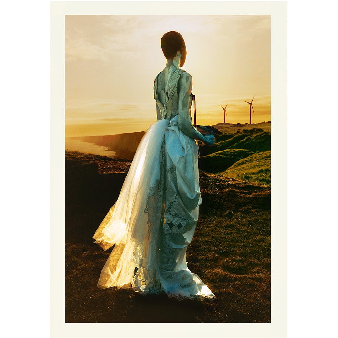 Woman in an ornate white gown, seen from the rear, standing on a grassy hill overlooking the coast at sunset, with distant wind turbines.