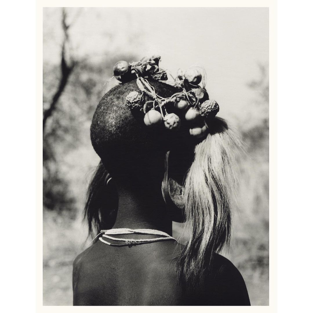 Back view of person with headdress made of fruit and feathers in black and white, outdoor setting, and shallow depth of field.