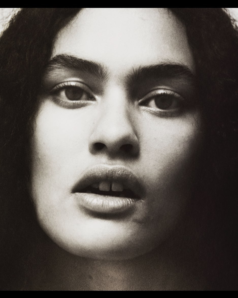 Close-up portrait of a woman with dark wavy hair, captured in a studio setting with soft lighting and a muted color palette.
