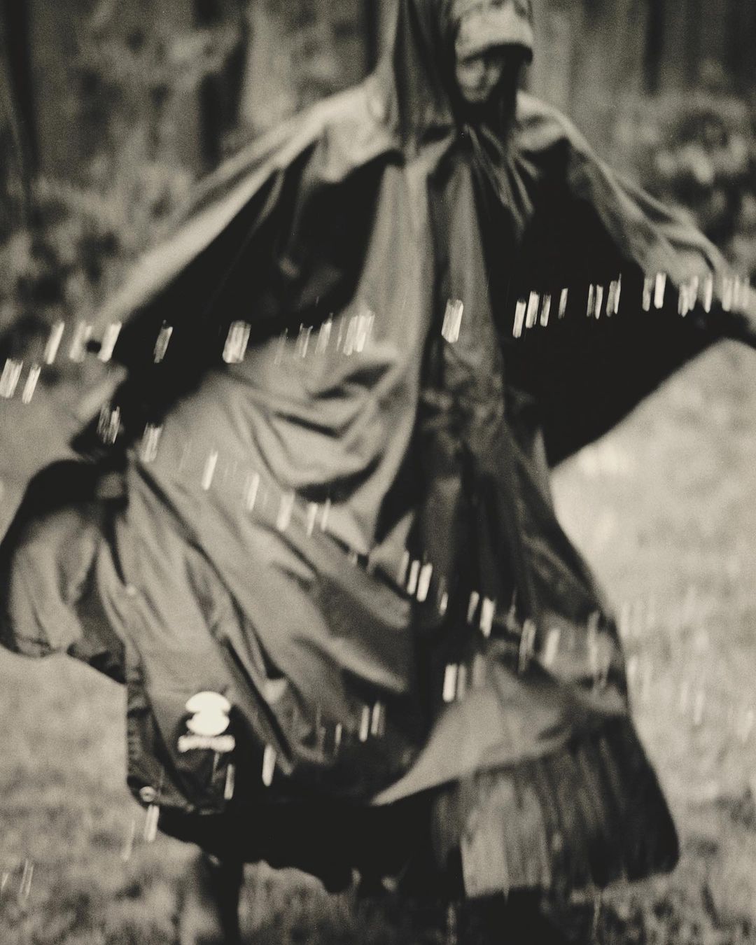 Person in a hooded poncho stands in a grassy field during rain. The scene is a black and white photograph, capturing a sense of motion.