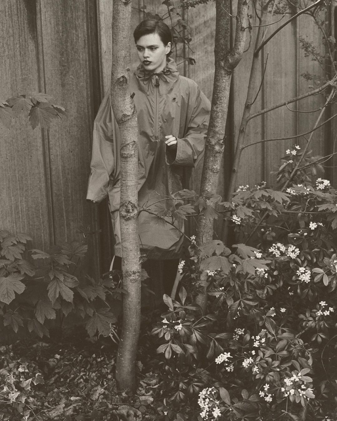 Woman in hooded raincoat poses outdoors amid trees and foliage, captured in a monochromatic photograph against a wooden fence.