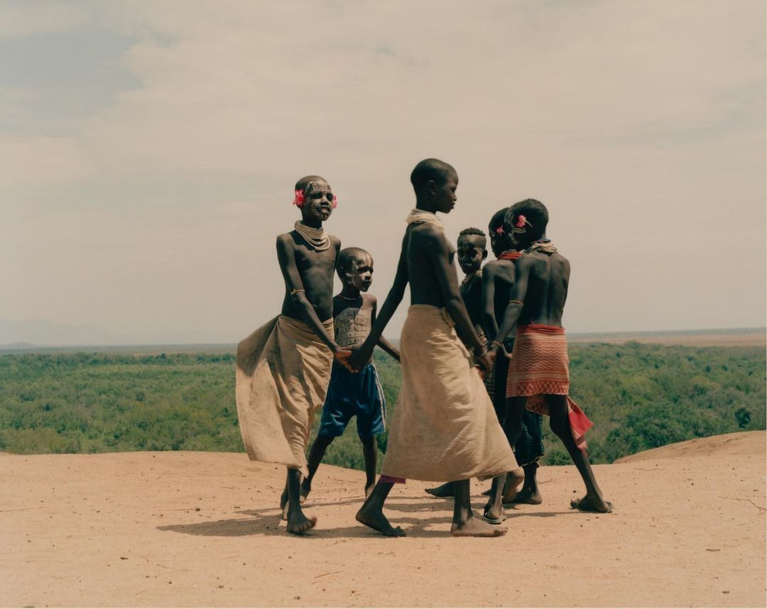 Group of six children outdoors in partial shade, wearing fabric wraps, head and neck decorations, on dirt near a forest backdrop.