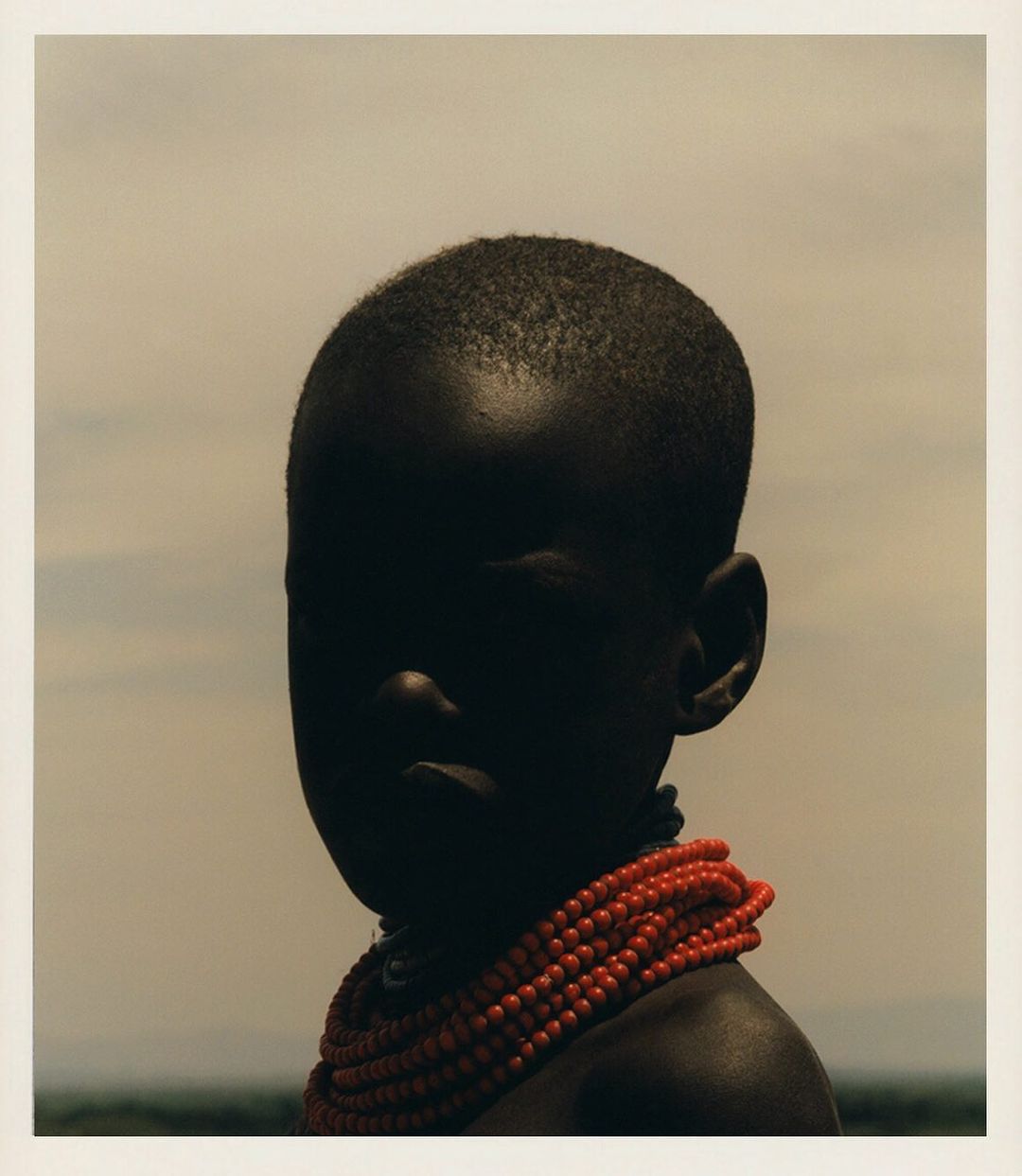 Portrait of child against a neutral background, wearing a coral-beaded necklace, with dramatic lighting that obscures facial features.
