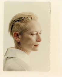 Profile portrait of Tilda Swinton with slicked-back blonde hair wearing a white collared shirt against a pale studio background.
