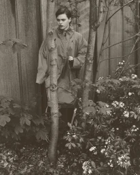 Model standing partially obscured behind tree trunks amidst leafy foliage in a monochromatic editorial portrait by Peter Jordanov