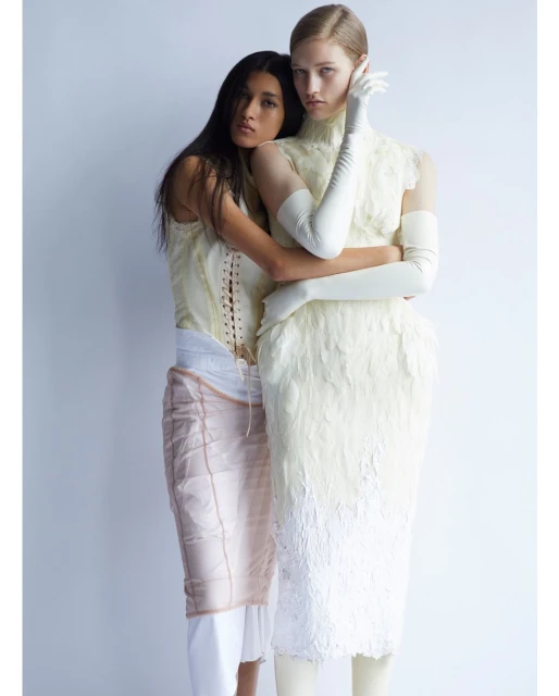 Two fashion models in a studio setting, one with dark hair embracing a blonde model wearing an elaborate cream-colored feather dress.