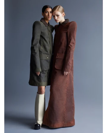 Two female models posing together against a grey studio backdrop, wearing structured long coats and tall boots.
