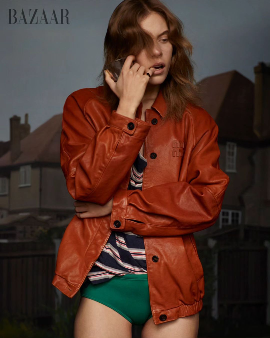 Editorial fashion shot of a female model in a brown leather jacket and green briefs, standing in front of a house at dusk.