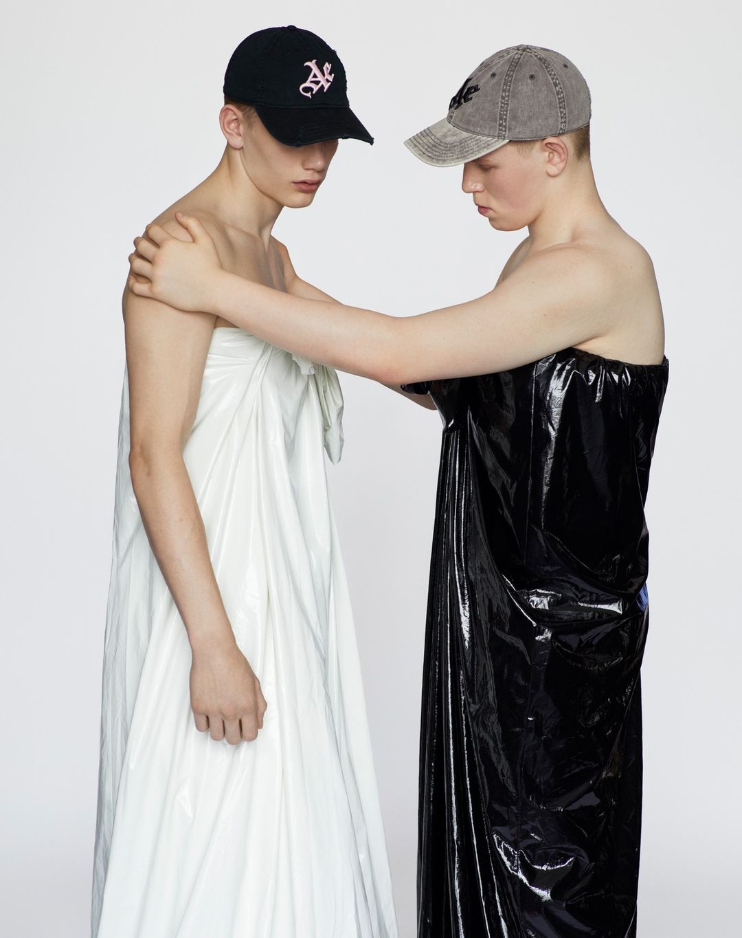Two young men in studio wearing caps, one in white fabric wrap, the other in a black plastic wrap. Fashion portrait.