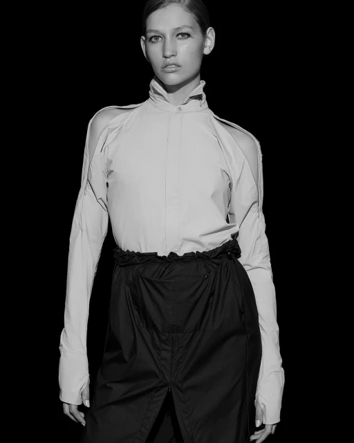 Monochrome studio portrait of a fashion model in a high-collared blouse and high-waisted skirt against a stark black background.