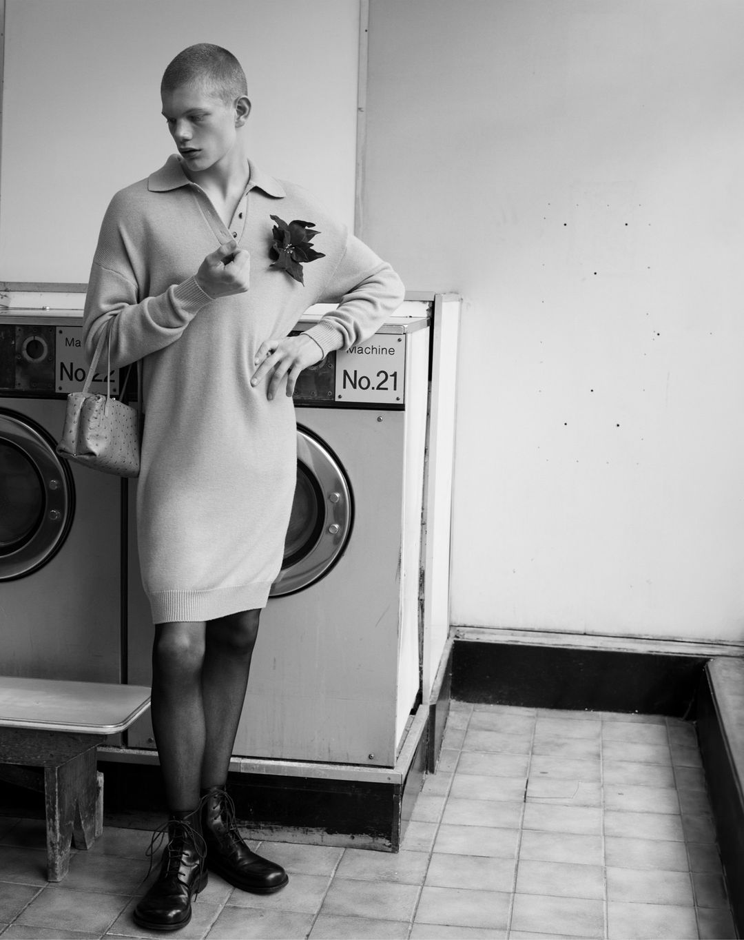 Black-and-white portrait of a model in a dress and tights, holding a flower in a laundromat, next to a row of washing machines.