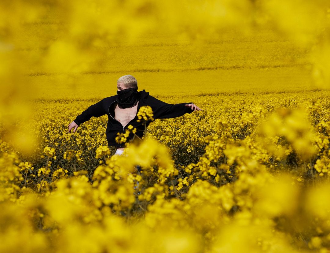 Person in black jacket stands in bright yellow field with arms outstretched, face covered by a black scarf.