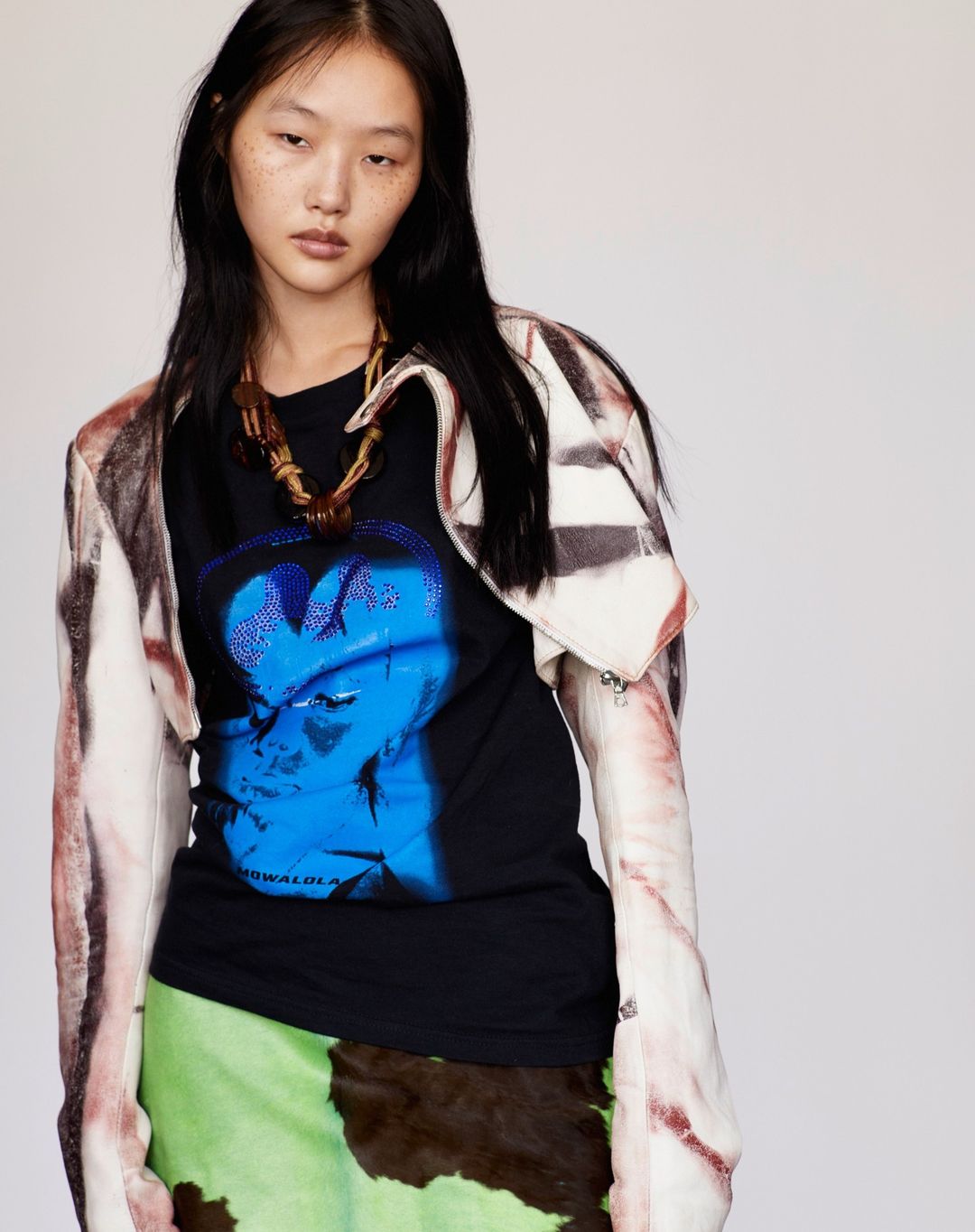 Portrait of an Asian woman in a black graphic tee, green skirt, and patterned jacket, standing in front of a neutral background.
