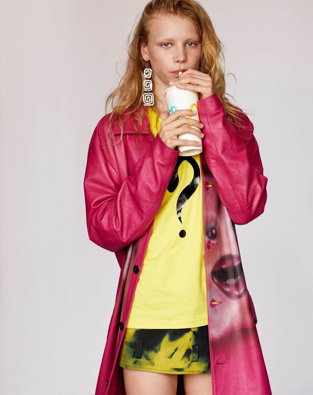 A blonde model in a pink coat sips from a McDonald's cup, wearing a yellow shirt with a question mark, against a white backdrop.