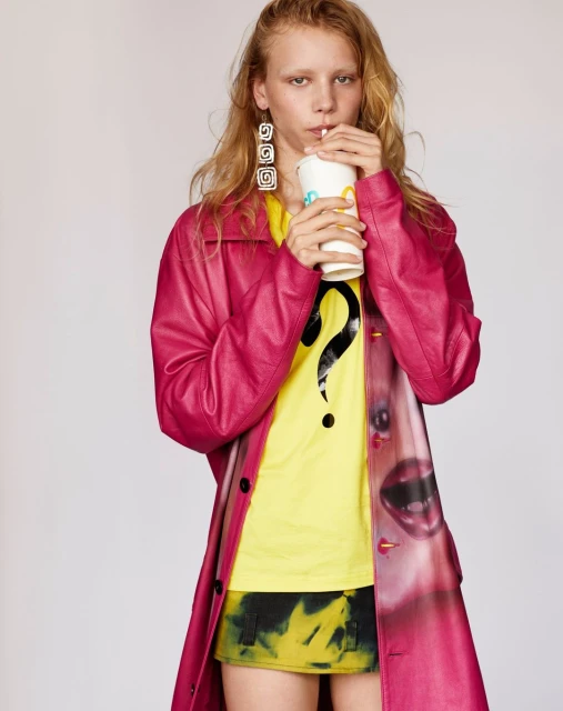 A blonde model in a pink coat sips from a McDonald's cup, wearing a yellow shirt with a question mark, against a white backdrop.