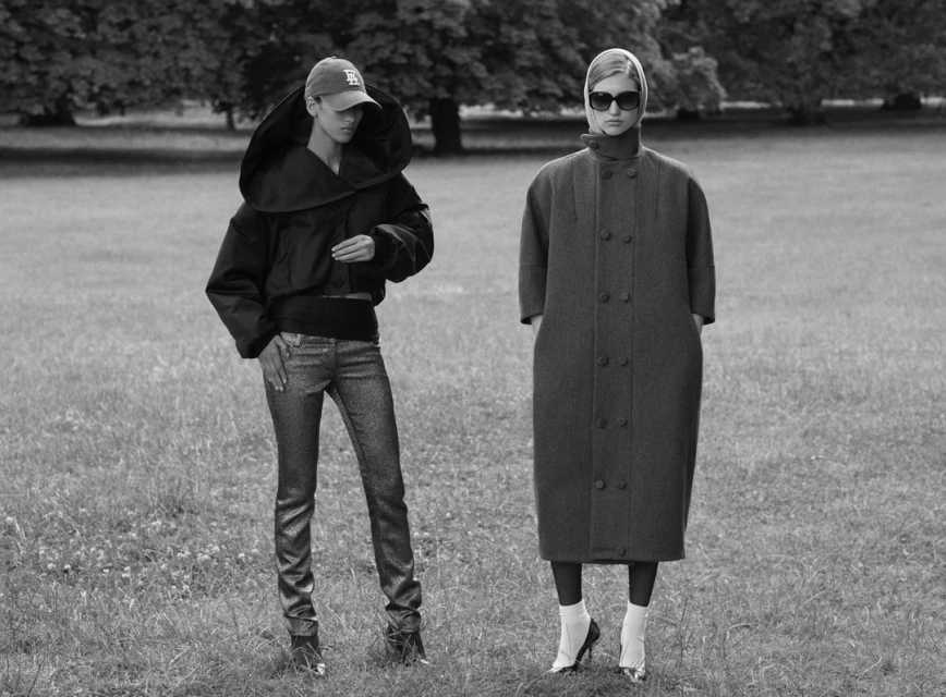 Two women in long coats and stylish outfits stand in a grassy field with trees in the background, showcasing fashion in a black and white photograph.