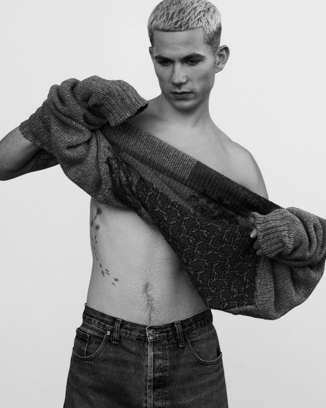 Monochrome studio portrait of a blonde man partially dressed in a grey knit sweater and blue jeans posing in front of a white backdrop.
