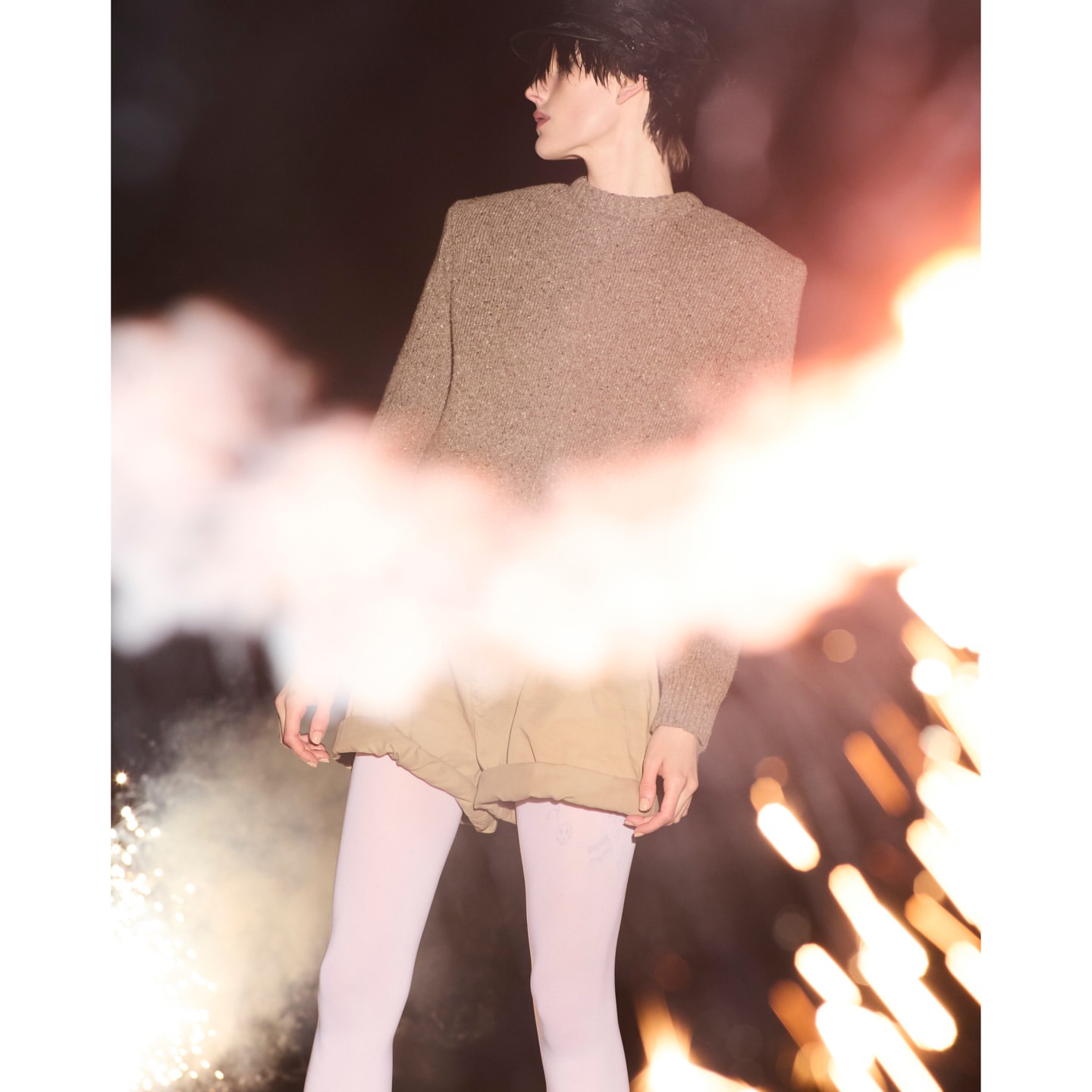 An adult female with fair skin, wearing a cap, sweater, and shorts, poses with smoke flares against a dark background in an outdoor setting.