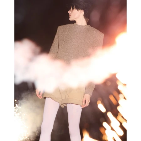 An adult female with fair skin, wearing a cap, sweater, and shorts, poses with smoke flares against a dark background in an outdoor setting.